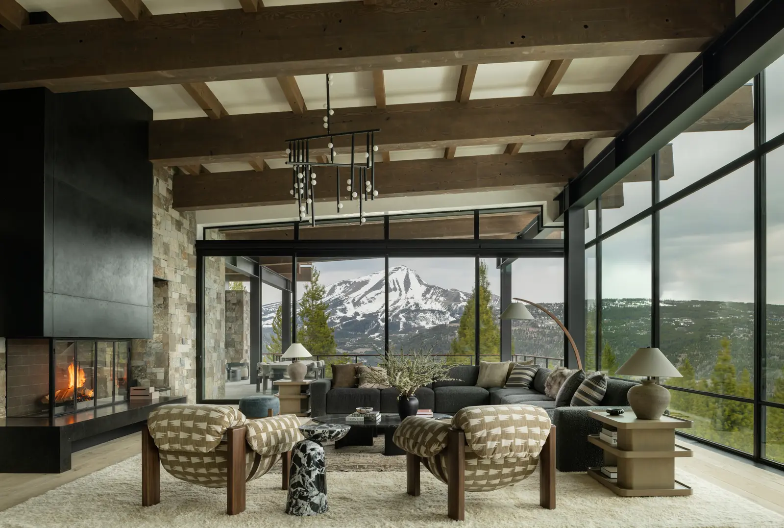 Great room under heavy timber beams, with a stone fireplace and walls of glass onto the mountains
